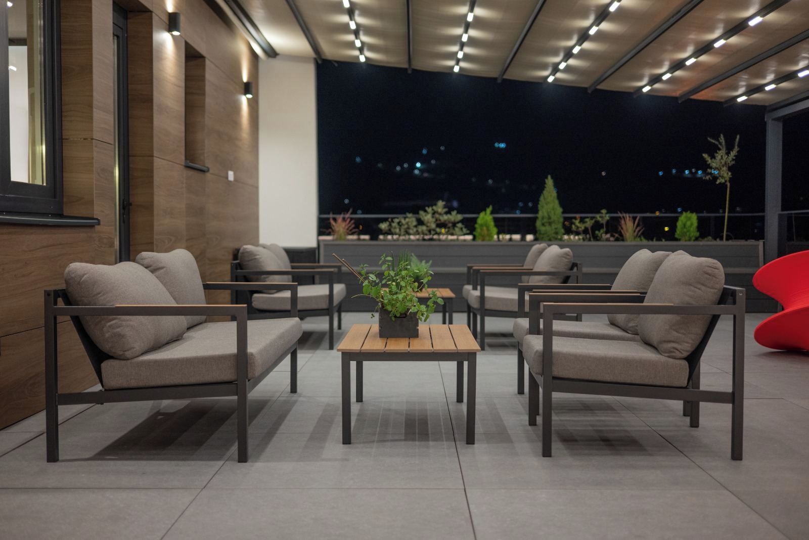 A modern outdoor seating area with sofas and a table, surrounded by plants and a view of the city at night.