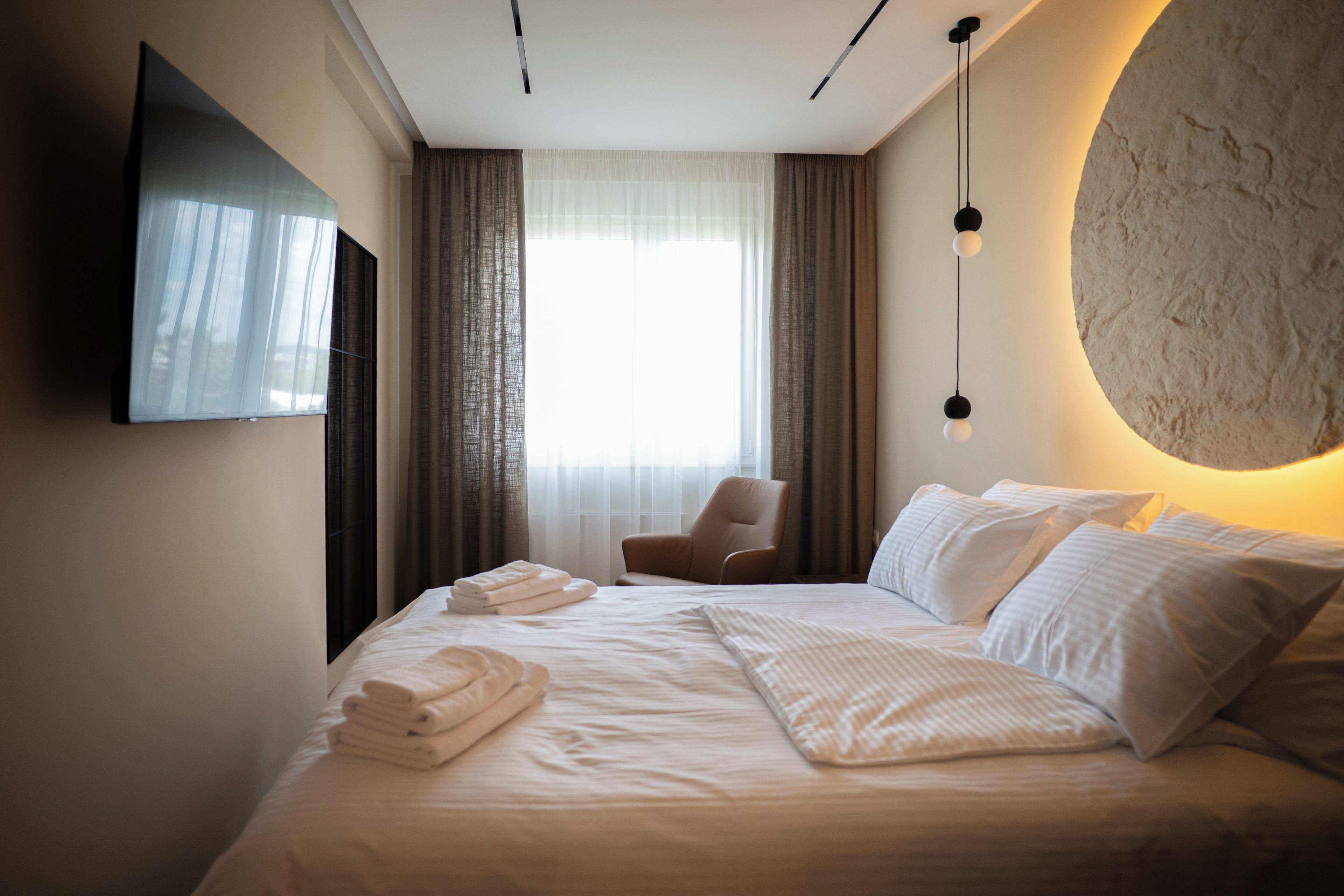 A hotel room with a bed, a chair, a TV mounted on the wall, and a large window with curtains.