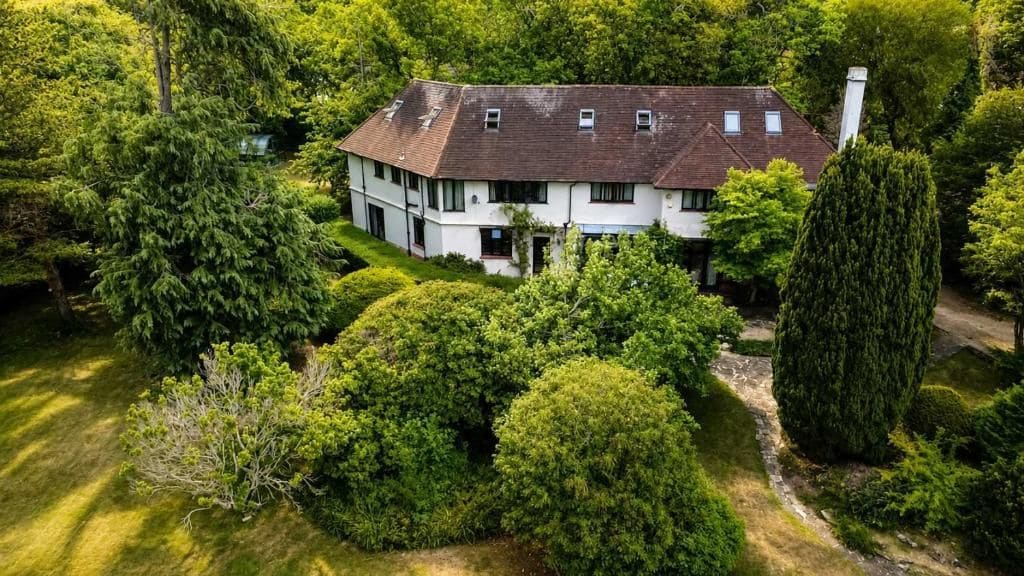 Una casa de dos pisos rodeada de abundante vegetación, con un camino de entrada visible y un árbol alto y delgado a la derecha.