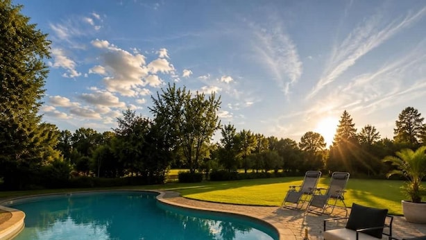A swimming pool with lounge chairs and a bottle of champagne, surrounded by trees and a clear sky. - Brook House (Stoke Poges)