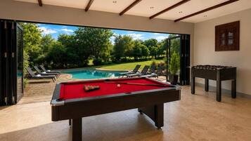 A pool table with a red felt surface, a foosball table, and a view of a swimming pool and greenery outside.