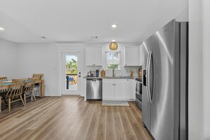 A modern kitchen with white cabinets, a stainless steel refrigerator, and a wooden dining table with chairs. - New 3BR Home- Less Than 1 Mile From The Beach (Gulfport)