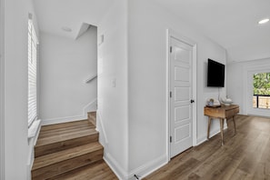 A staircase with wooden steps leading to a white door, a flat-screen TV mounted on the wall, and a small wooden table with a bowl and a sign. - New 3BR Home- Less Than 1 Mile From The Beach (Gulfport)