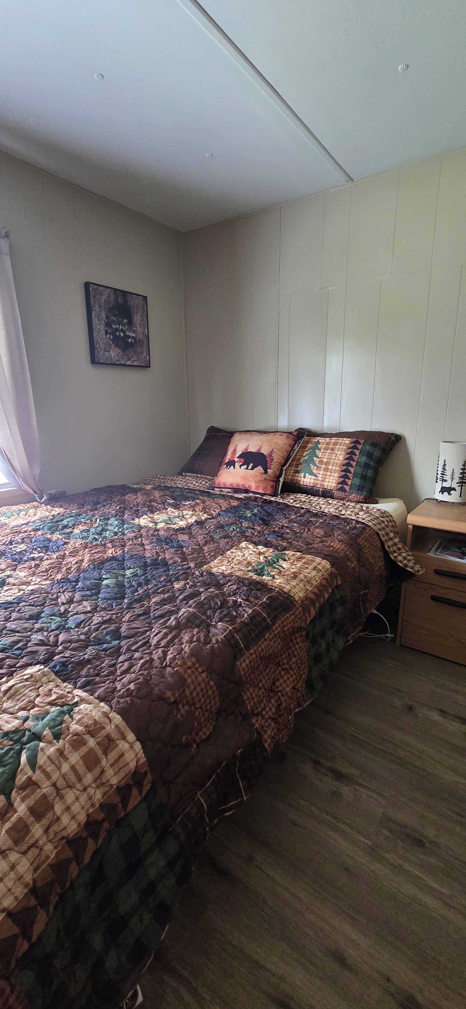 A bedroom with a large bed, a nightstand, a wall-mounted shelf, and a window with curtains.