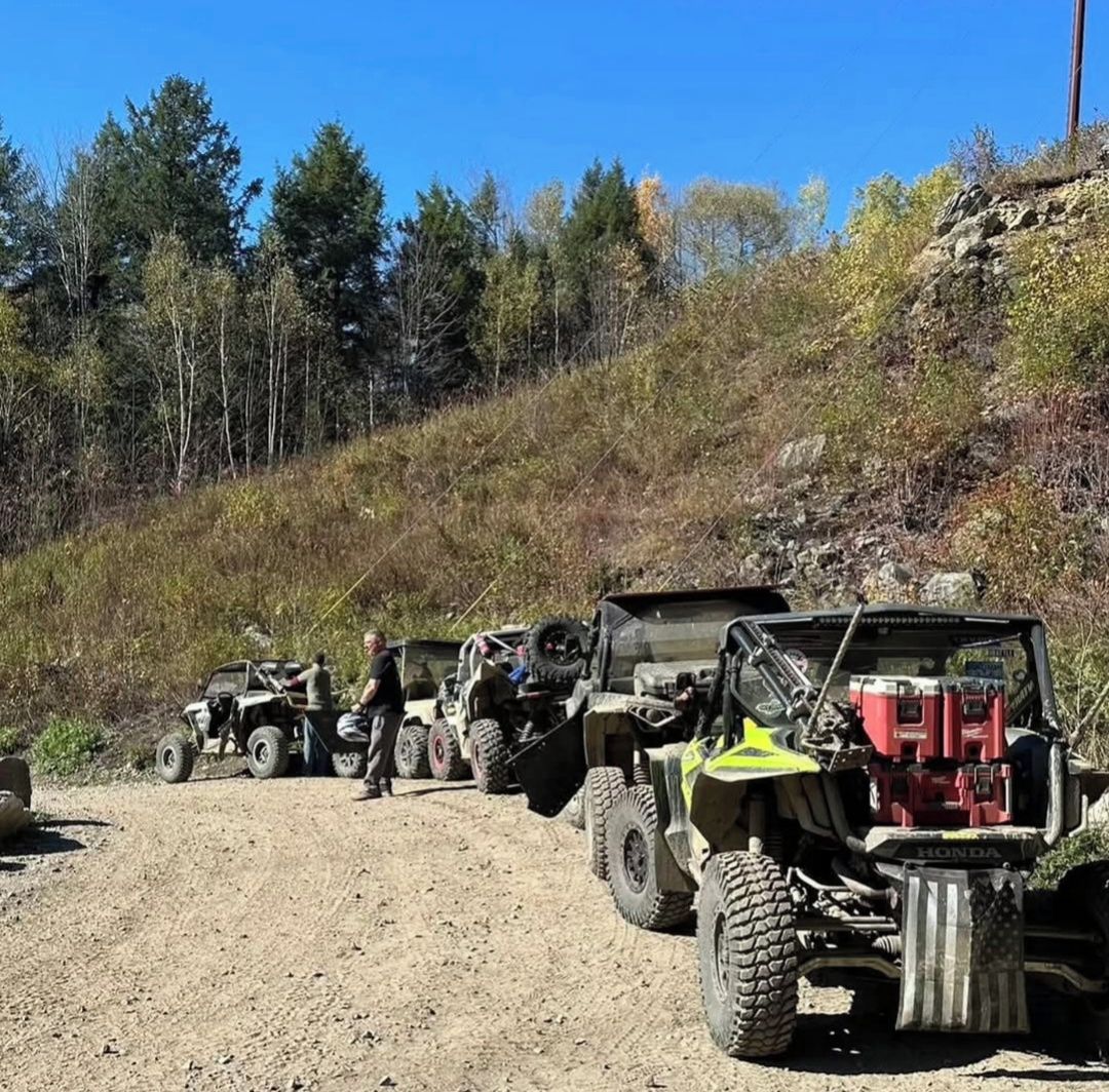 Un groupe de véhicules tout-terrain est garé sur un chemin de terre, avec une colline boisée en arrière-plan.