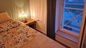 Un dormitorio con cama, lámpara de noche, ventana con vista a la calle y cortinas.