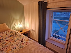 Ett sovrum med en säng, en sänglampa, ett fönster med utsikt över gatan och gardiner.