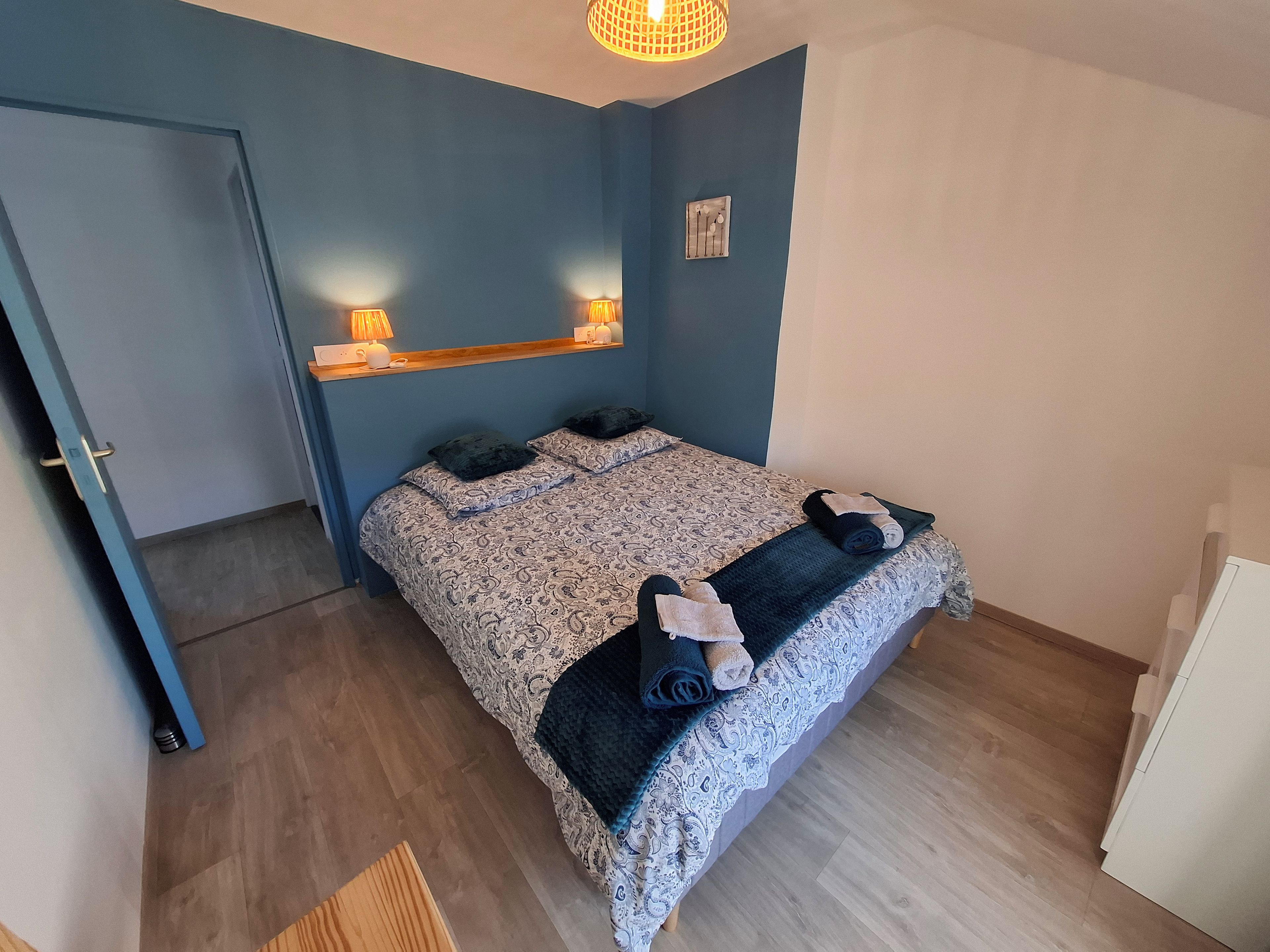 Un dormitorio con cama, una mesita de noche de madera, un estante en la pared con lámparas y una puerta.