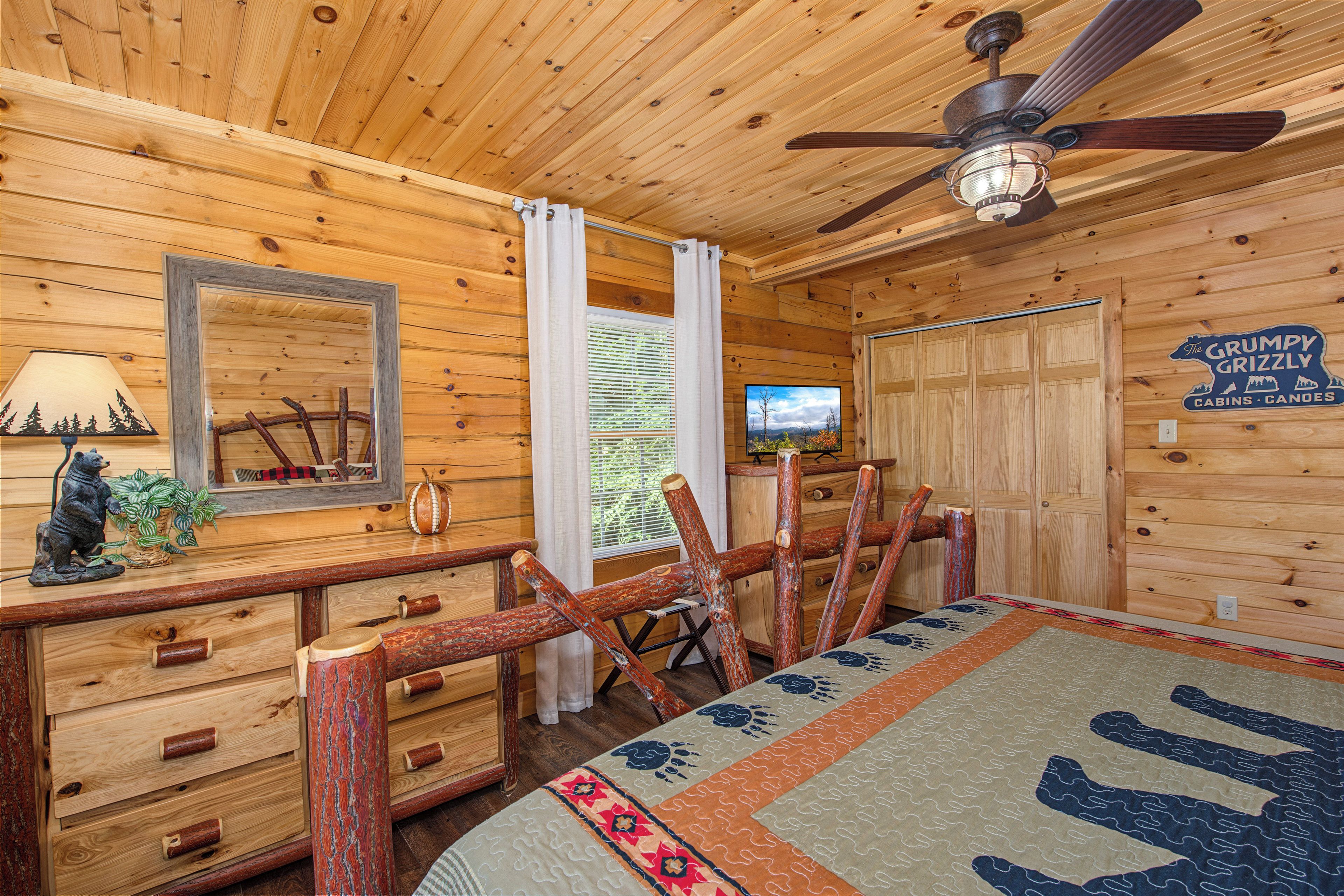 A cabin interior with wooden walls, a ceiling fan, a bed with a patterned bedspread, a dresser with wooden handles, a mirror, and a television on a wooden cabinet.