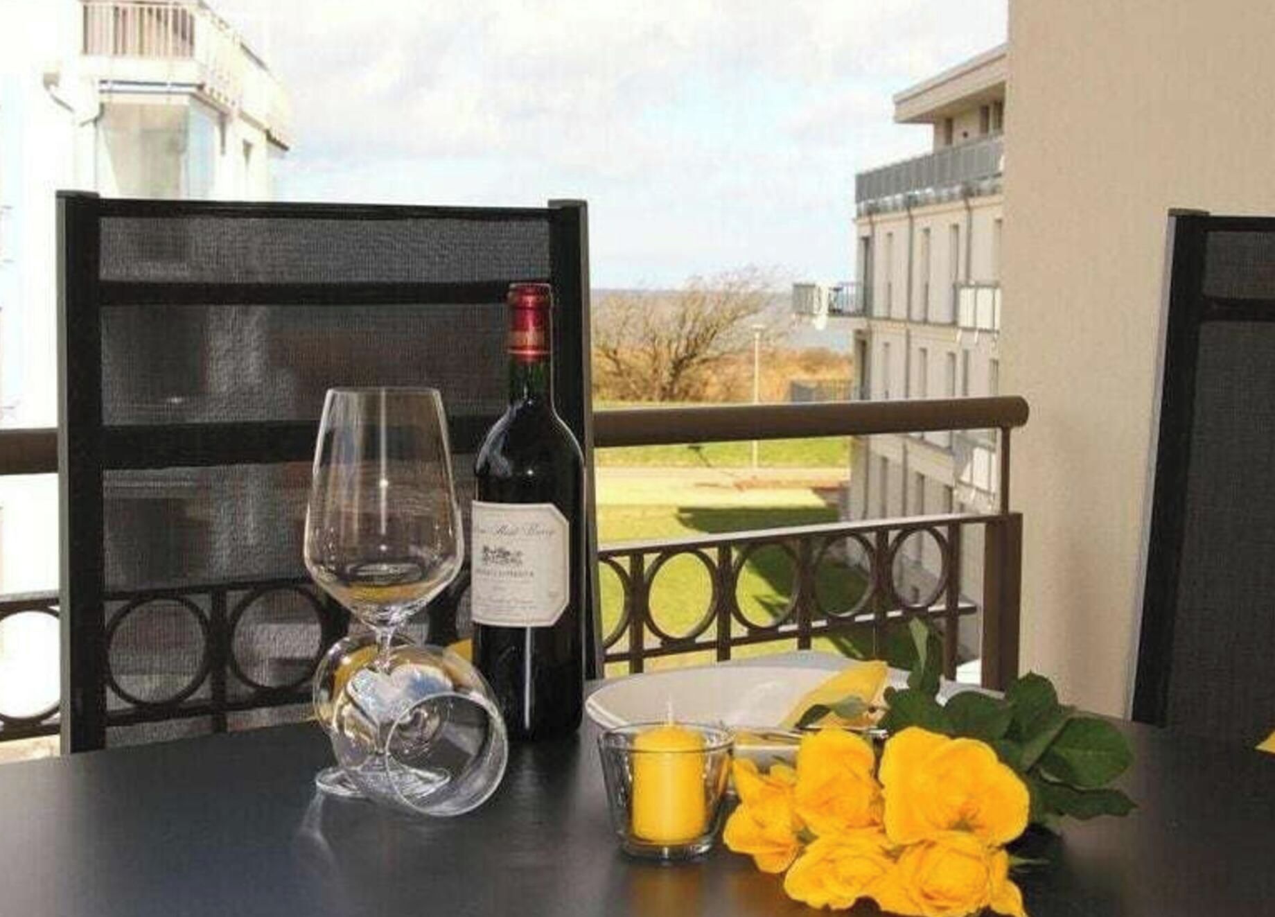 A table with a wine bottle, a wine glass, a candle, and yellow flowers on a balcony overlooking a building.