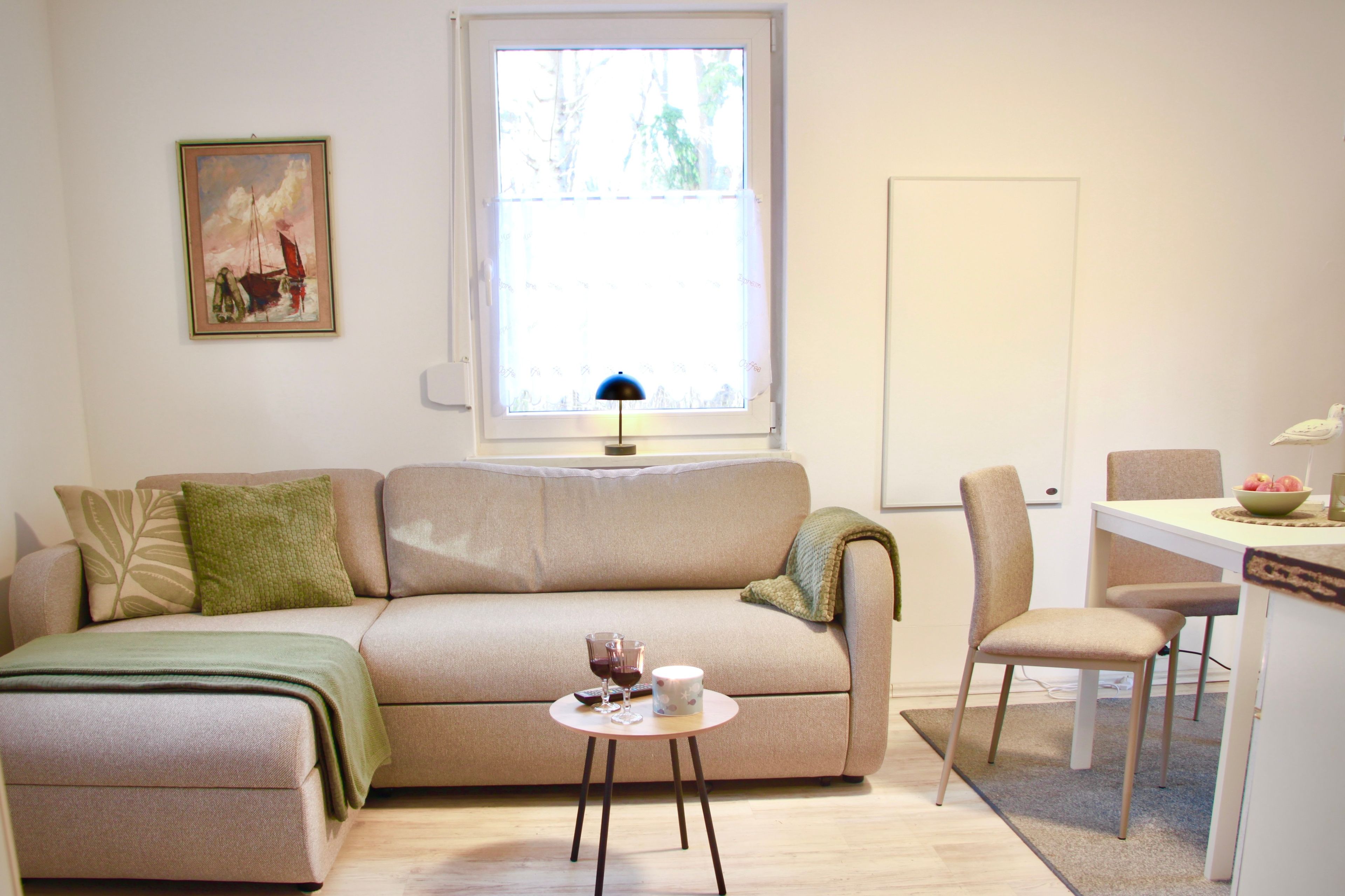 A living room with a sofa, a small table with drinks, a chair, a window with a view, and a painting on the wall.