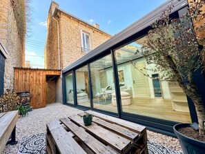 A modern outdoor seating area with a wooden bench and a potted plant, a glass wall, and a view of an interior space with furniture. - Dinan • Renovated 2-bedroom house & exterior • Train station and downtown within walking distance (Dinan)