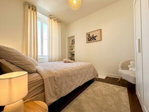 A bedroom with a bed, bedside lamp, and a wall-mounted shelf with decorative items. - Dinan • Renovated 2-bedroom house & exterior • Train station and downtown within walking distance (Dinan)