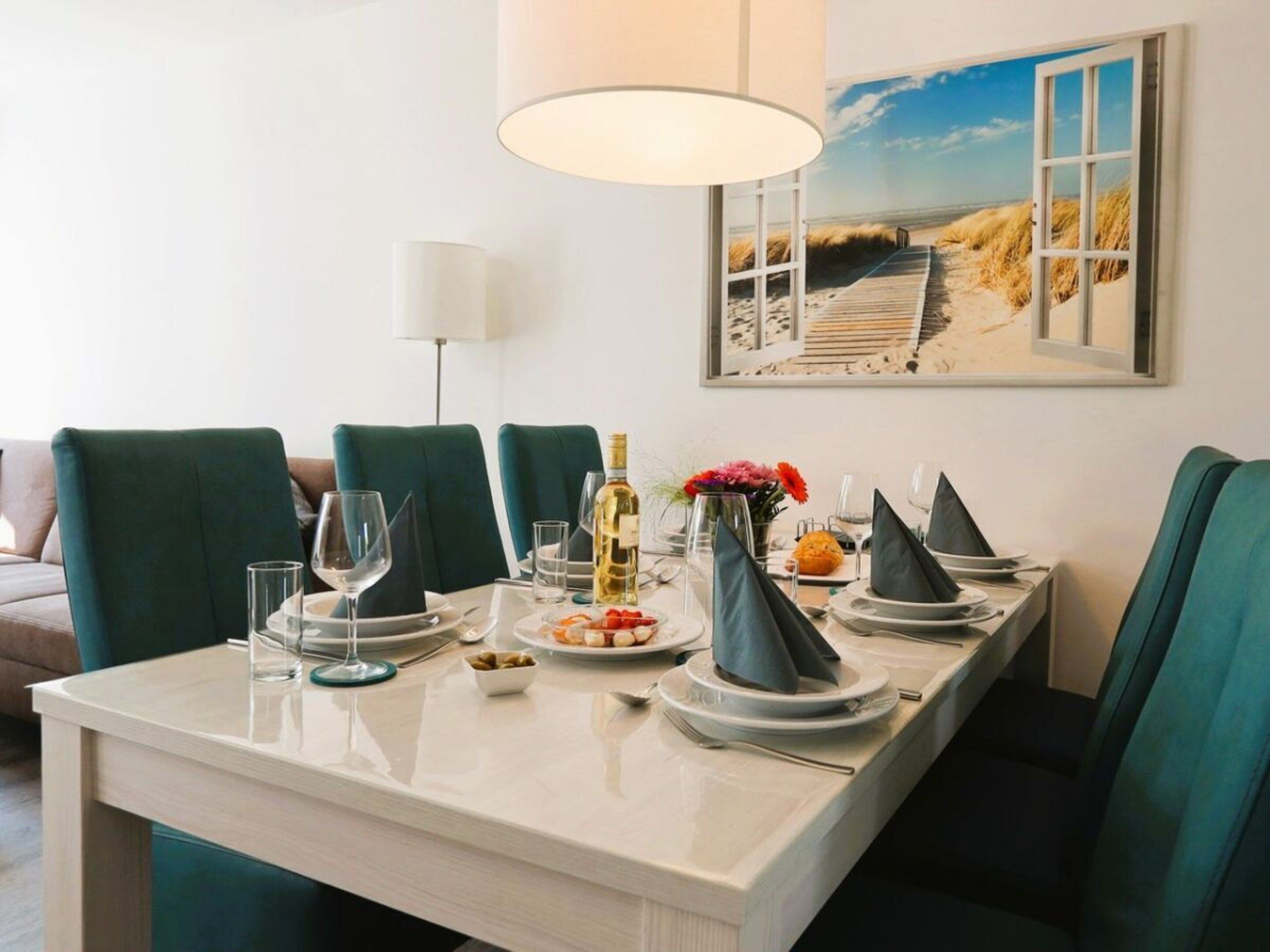 Une table à manger dressée pour six personnes, avec un tableau représentant un paysage côtier accroché au-dessus.