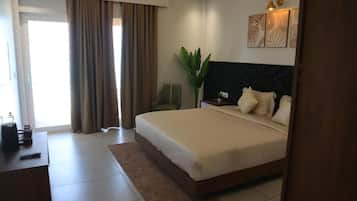 A hotel room with a large bed, a desk with a chair, a potted plant, and artwork on the wall.