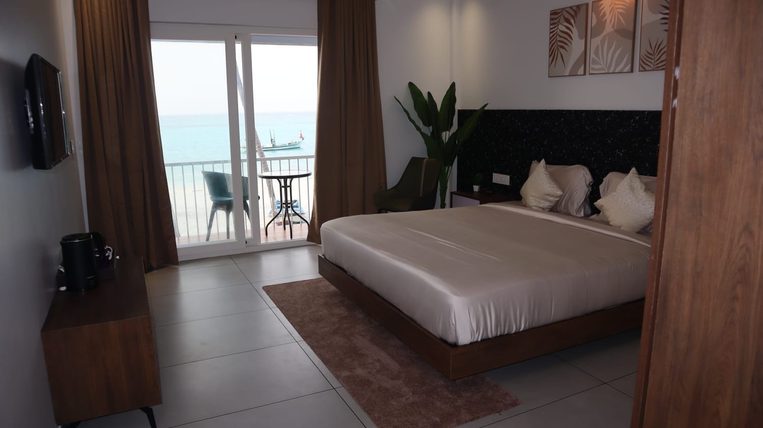 A hotel room with a bed, a TV, a coffee maker, a plant, and a view of the sea.