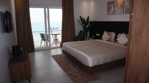 A hotel room with a bed, a TV, a coffee maker, a plant, and a view of the sea.
