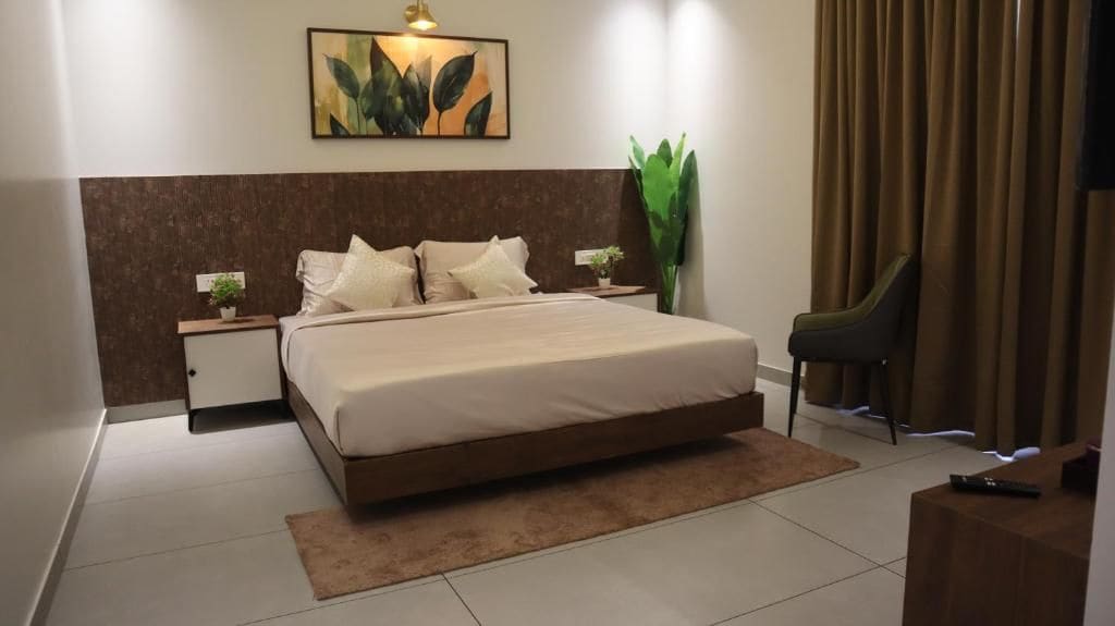 A hotel room with a bed, bedside tables, a nightstand, a chair, a large plant, and a painting on the wall.