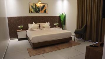 A hotel room with a bed, bedside tables, a nightstand, a chair, a large plant, and a painting on the wall.
