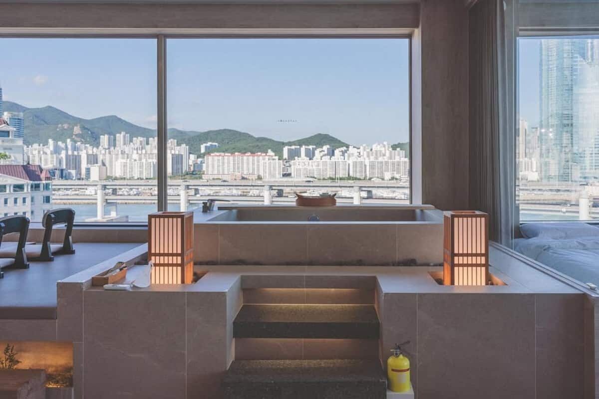 Ein modernes Hotelzimmer mit großem Fenster und Blick auf die Stadt, einer kleinen Sitzecke und einer Treppe, die in eine obere Ebene führt.