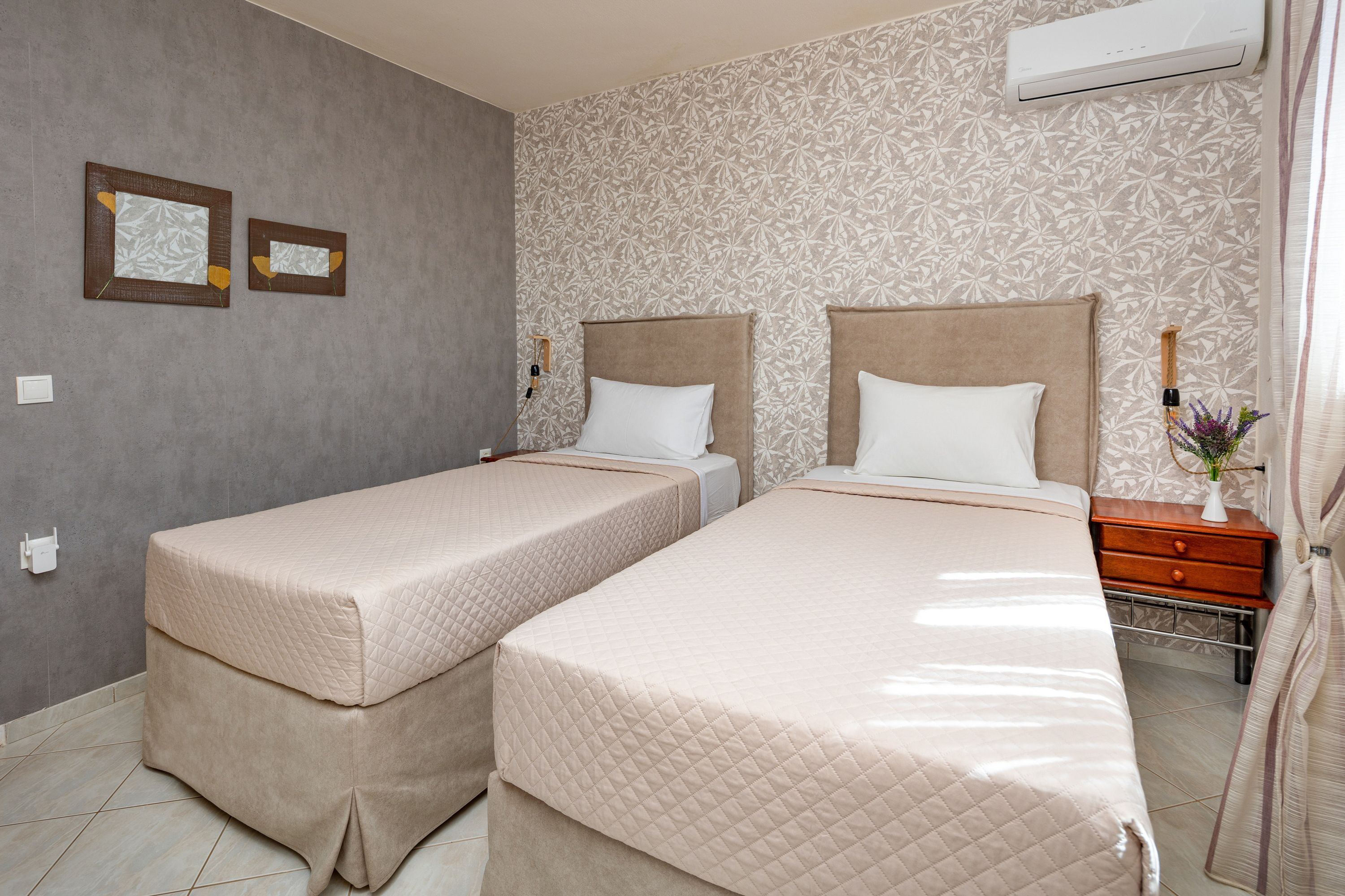 A hotel room with two beds, a wall-mounted air conditioner, and a small bedside table with a vase of flowers.
