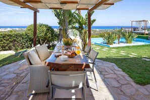 Eine Terrasse mit Frühstückstisch, mit Blick auf einen Pool und das Meer.