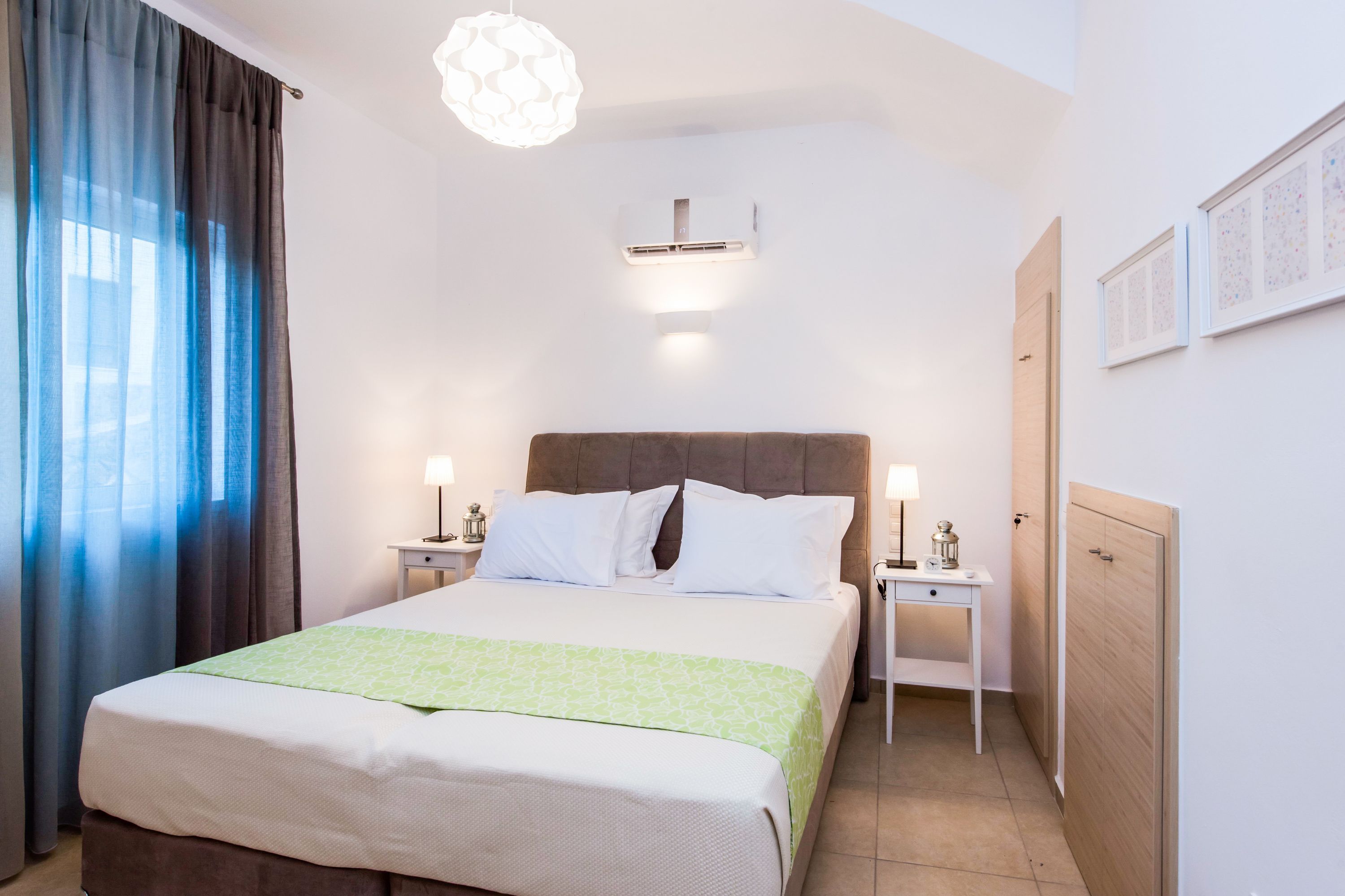 Habitación de hotel con cama, mesitas de noche, lámpara, aire acondicionado instalado en la pared y una puerta.