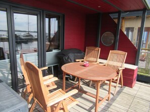 Eine Terrasse im Freien mit einem runden Holztisch, vier Stühlen, einem Grill und Blick auf ein Gewässer durch große Fenster.