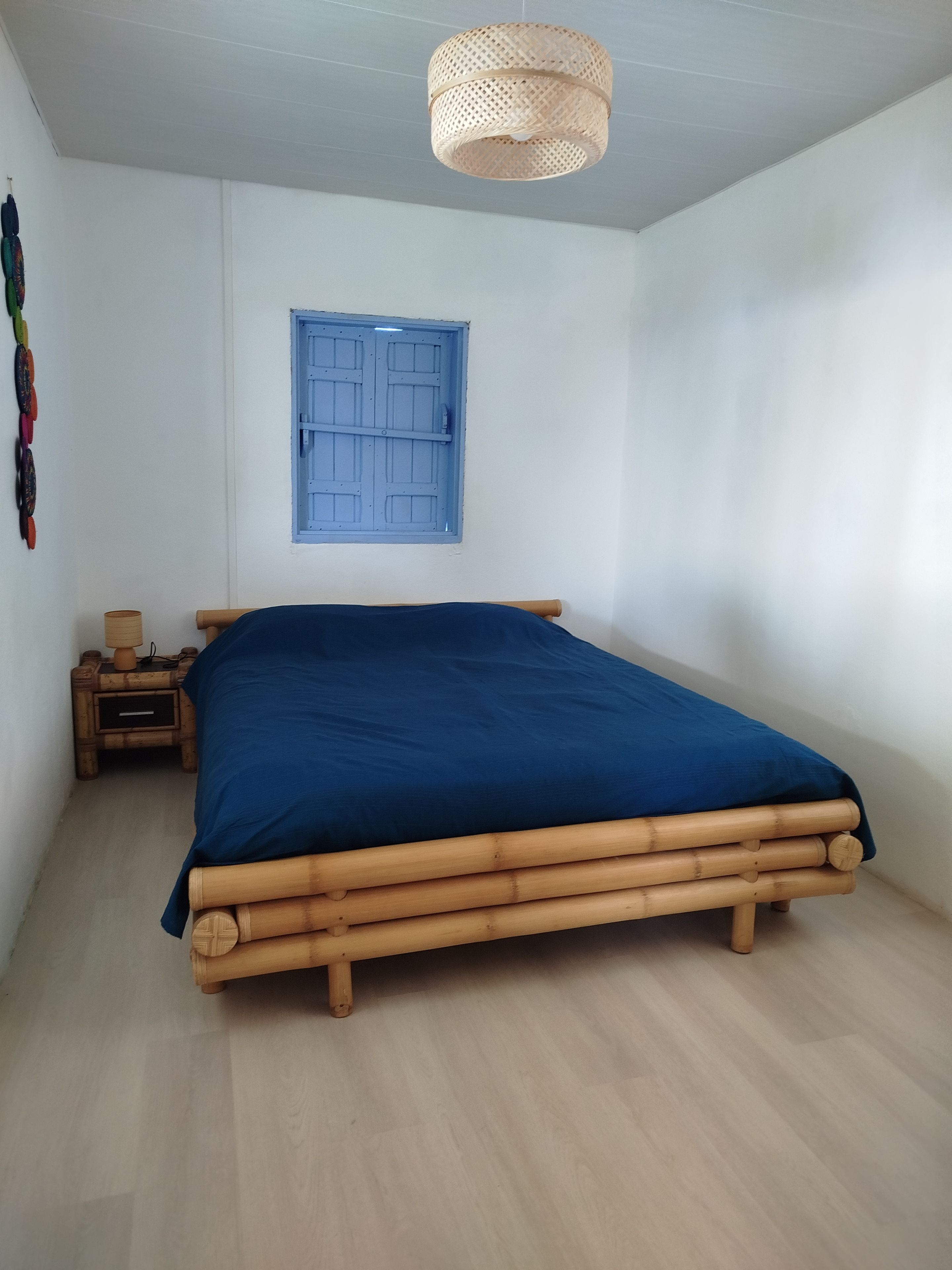 Une chambre avec un lit, une fenêtre avec des volets bleus, un luminaire fixé au mur et une petite table de chevet en bois avec une lampe.