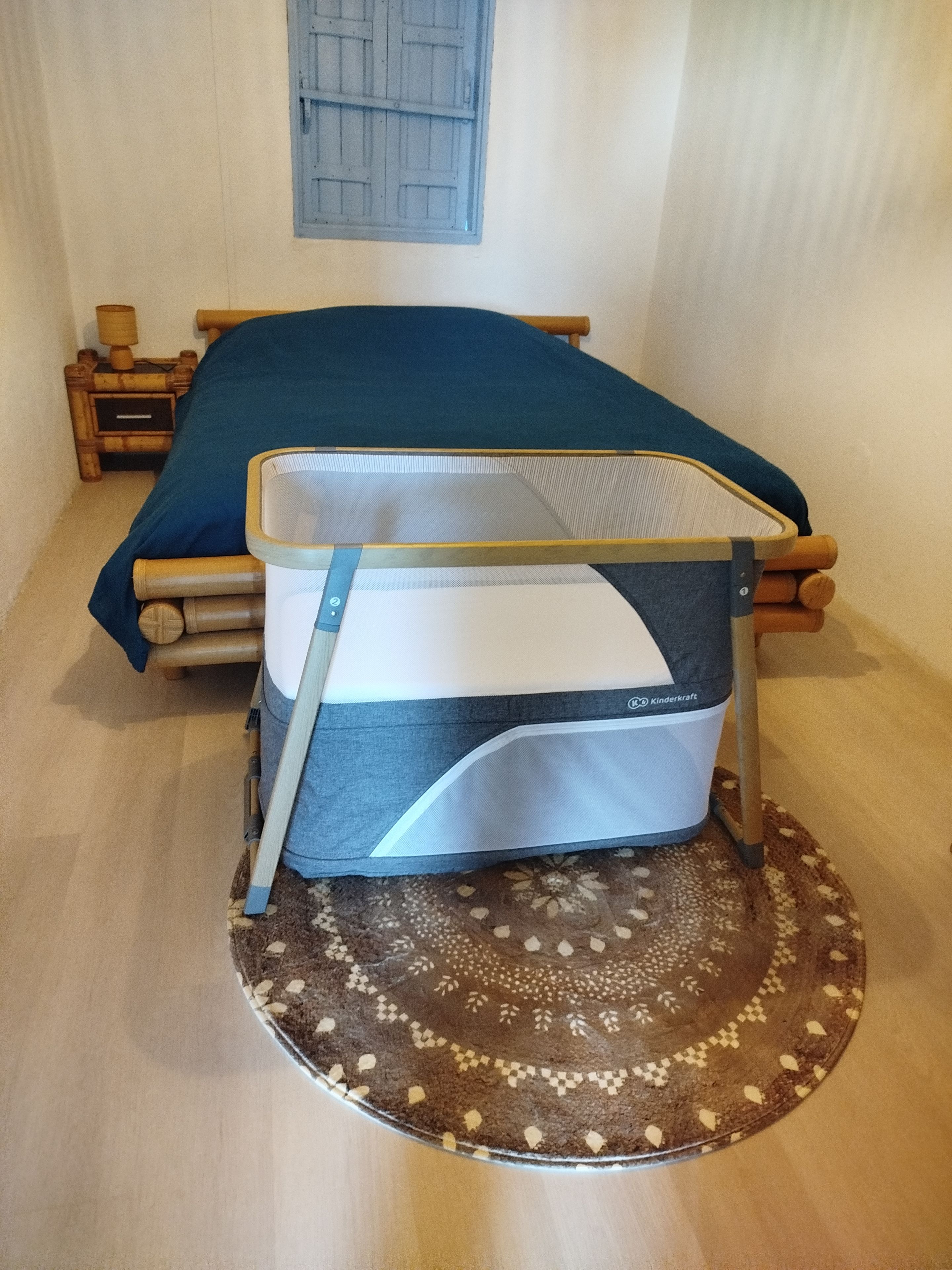 Ein Bett mit einer Matratze mitten im Raum, ein Nachttisch aus Holz mit einer Lampe und ein Fenster mit blauem Rollo.