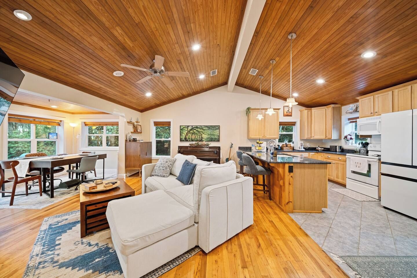 A spacious living area with a wooden ceiling, a dining table, a sofa, and a kitchen with wooden cabinets and a white refrigerator.