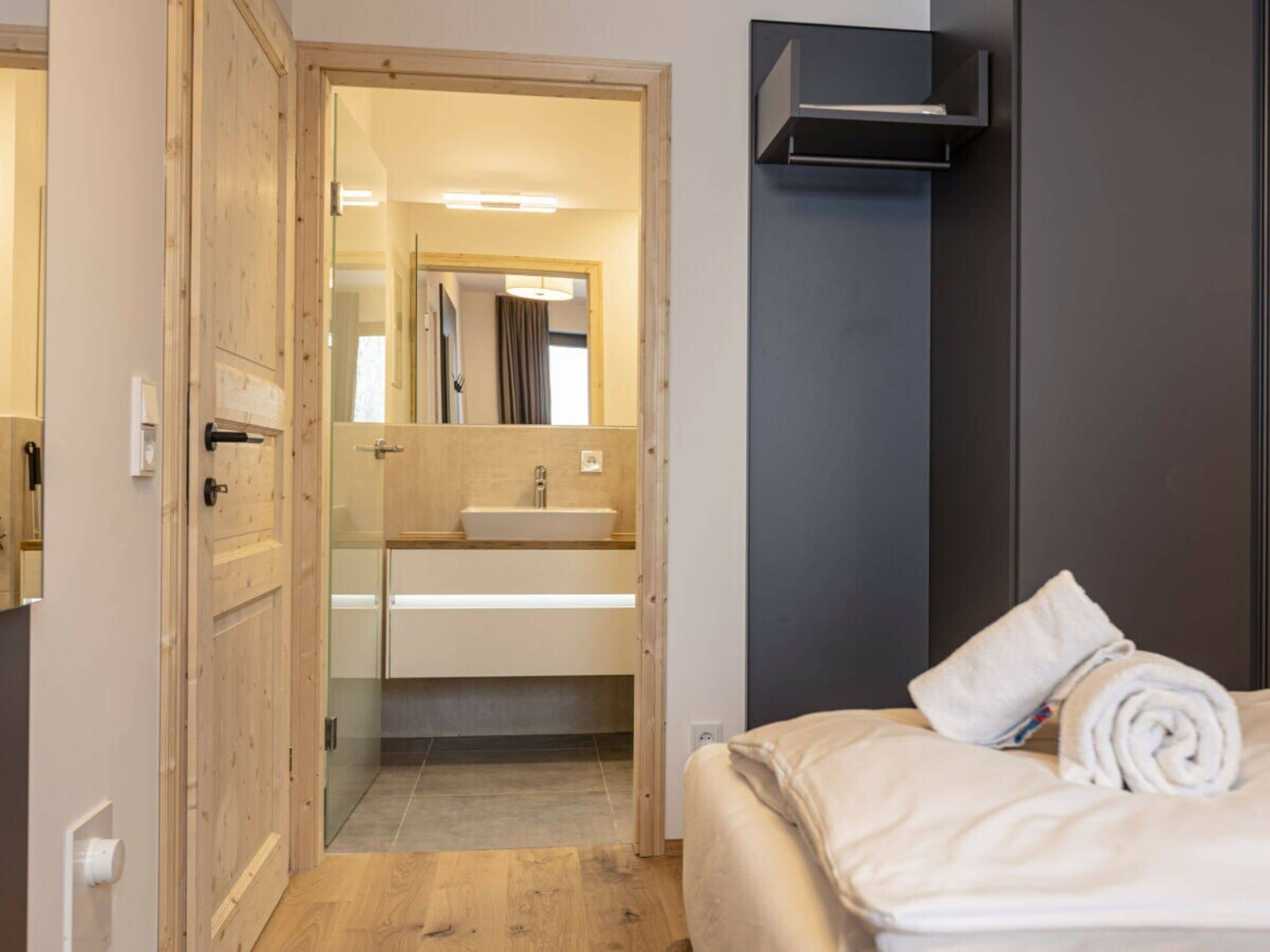A hotel room with a wooden door, a bathroom with a sink and mirror, and a bed with a rolled towel.