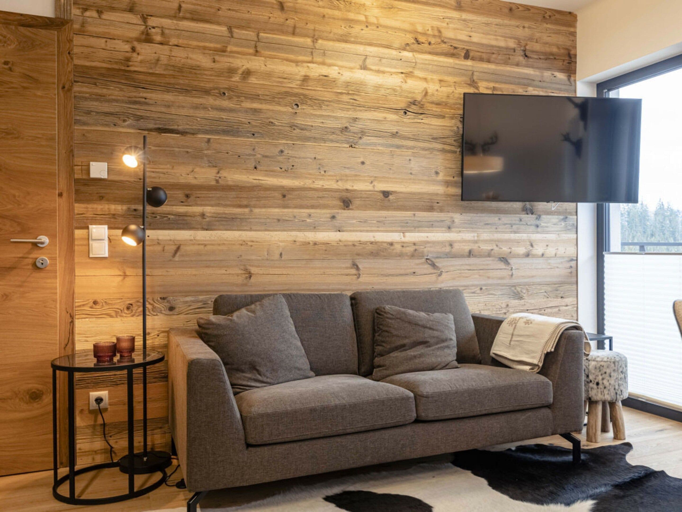 Ein modernes Wohnzimmer mit einer Holzvertäfelung, einem grauen Sofa, einer schwarzen Stehlampe und einem an der Wand befestigten Fernseher.