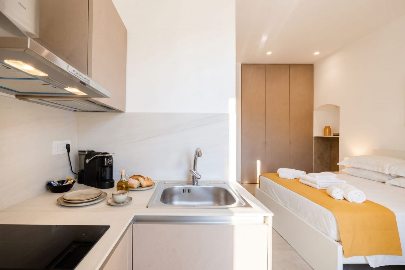 Una habitación de hotel compacta con kitchenette, una cama con manta amarilla y una mesita de noche con toallas.