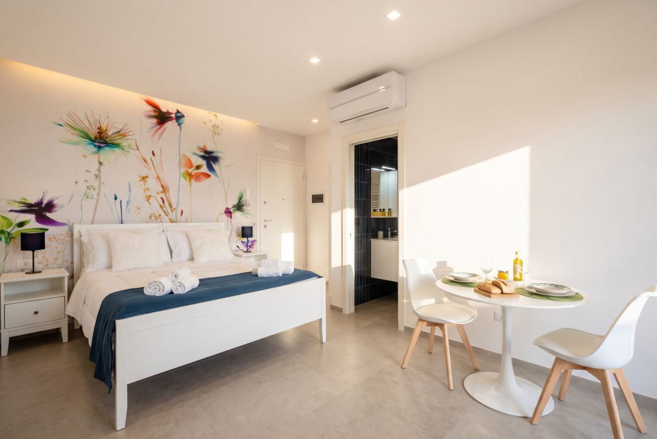 Een moderne slaapkamer met een bed, nachtkastjes, een zithoek met stoelen en een keuken op de achtergrond.