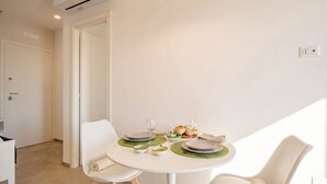 En spiseplads med et rundt bord dækket til to personer, en hvid stol, et lille bord med brød og et glas, samt en vægmonteret lampe.