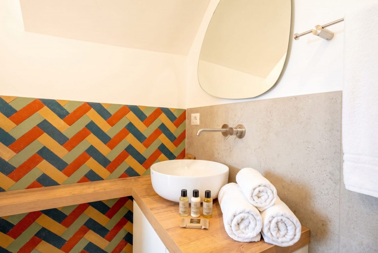 Une salle de bain avec un mur recouvert de carreaux disposés en motif chevrons, un lavabo blanc, un miroir et des serviettes soigneusement pliées.