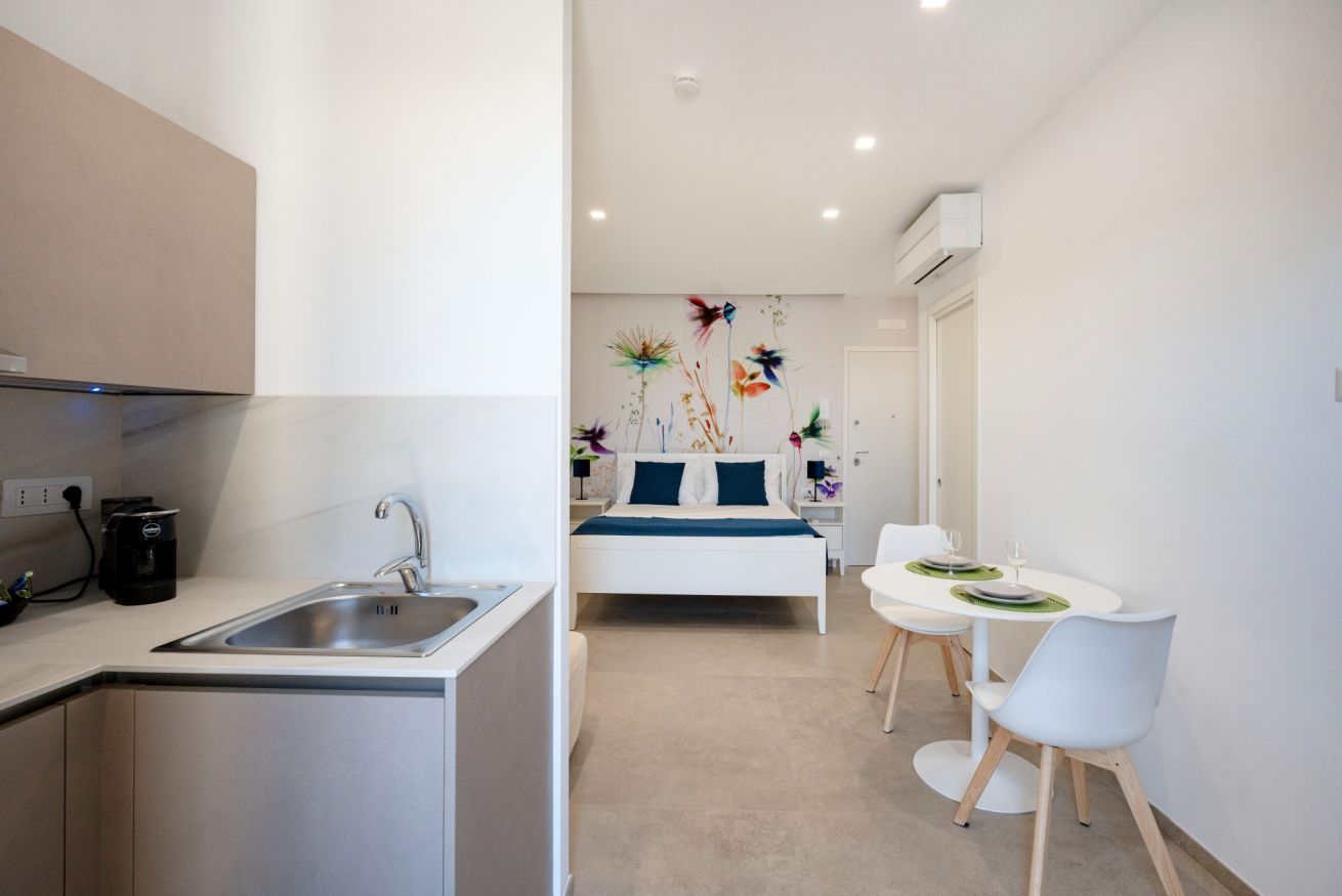 Una moderna camera d'albergo con angolo cottura, un letto con testiera blu, una zona pranzo con tavolo e sedie, e una parete decorata con motivi floreali.