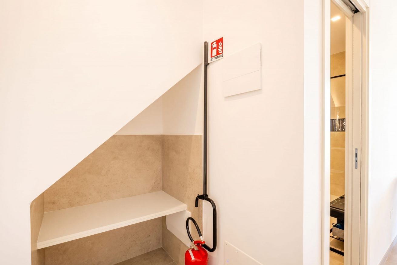 Un coin de pièce avec une étagère blanche, un extincteur fixé au mur et une porte donnant sur une autre pièce.