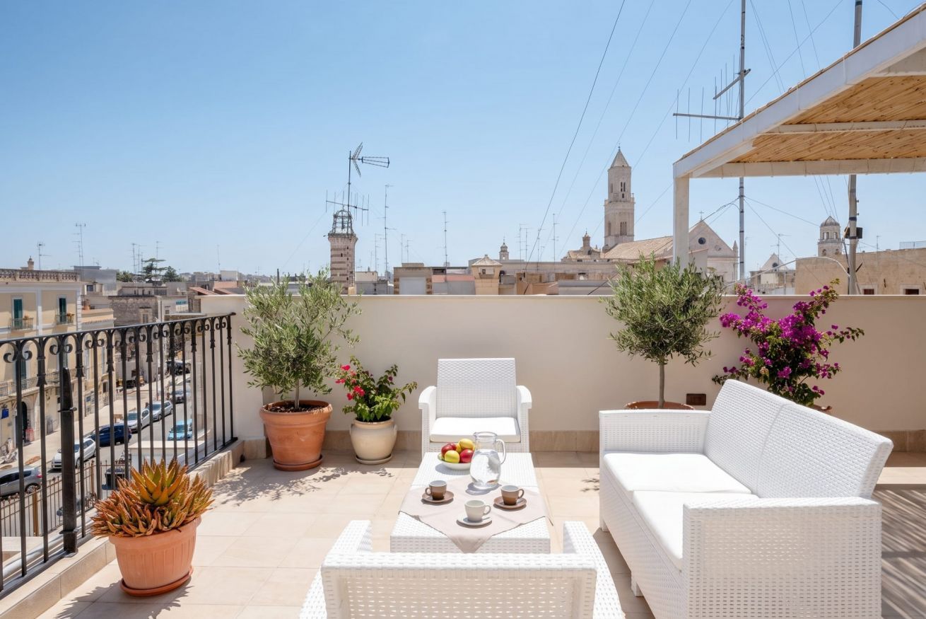 Una terrazza sul tetto con un divano in vimini bianco, un tavolino con una brocca e tazze, piante in vaso e vista sulla città.