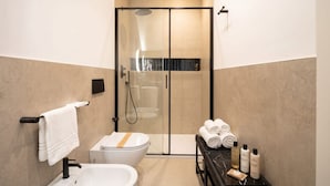 Et moderne badeværelse med en brusekabine i glas, en hvid vask og et toilet. Håndklæder og toiletartikler er placeret på en hylde.