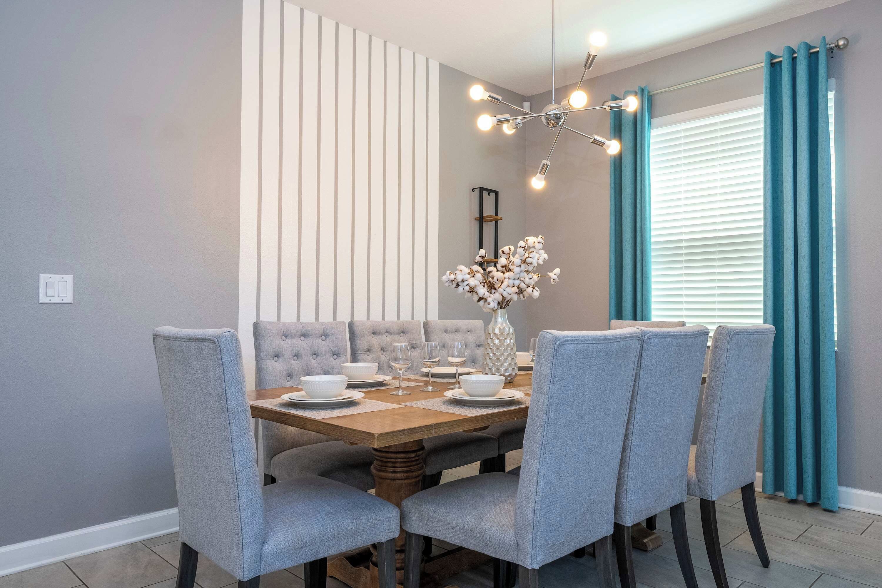 Comedor con mesa de madera, sillas tapizadas en gris, un florero con flores y una lámpara moderna.
