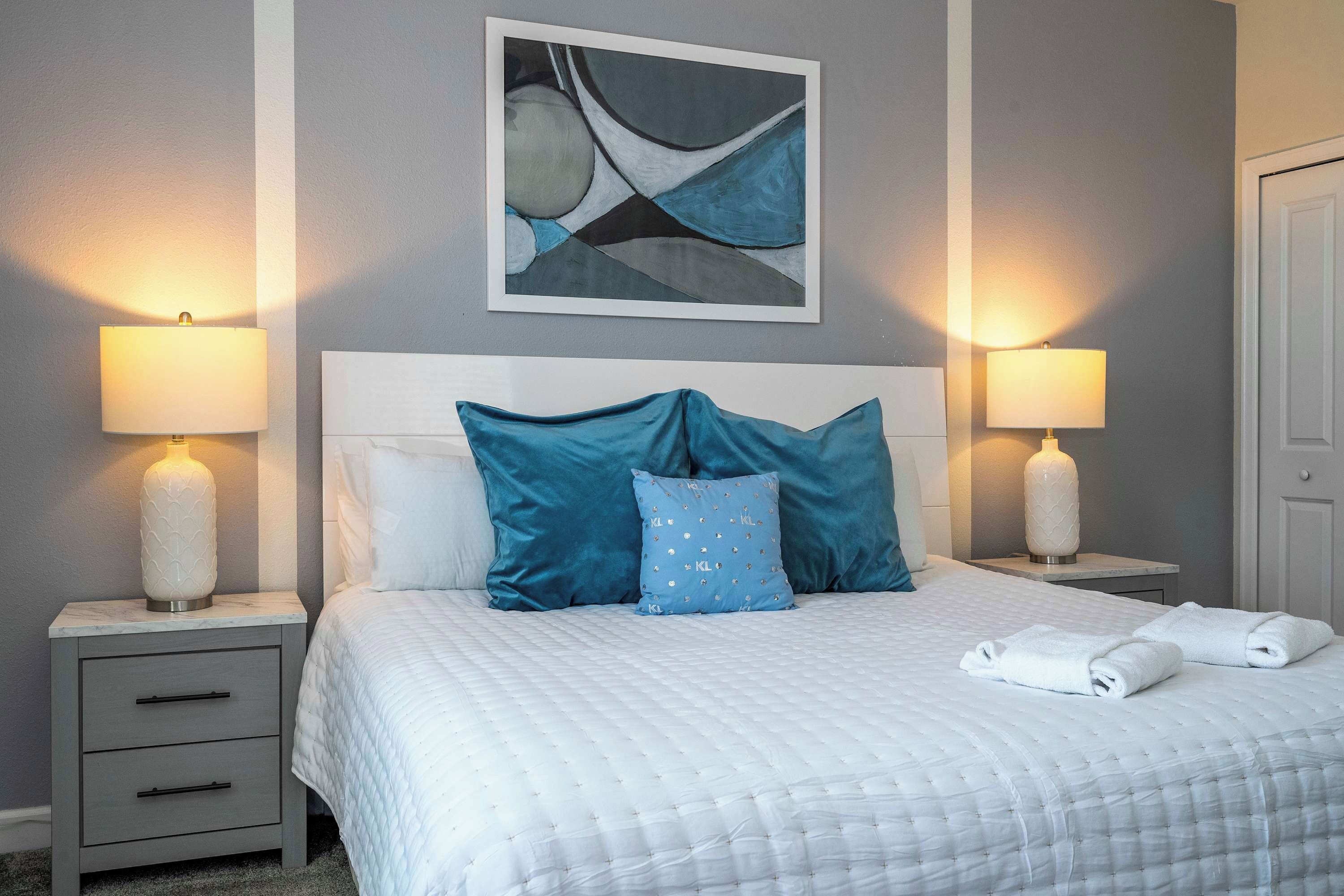 Un lit bien fait, avec une couette blanche et des oreillers bleus, flanqué de deux tables de chevet sur lesquelles reposent des lampes, dans une pièce dont un mur est gris et au-dessus du lit, on peut voir un tableau abstrait encadré.