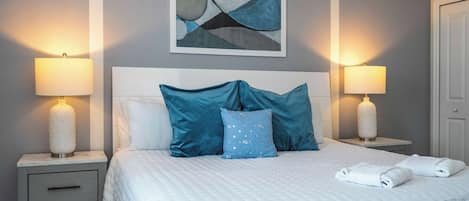 Un lit bien fait, avec une couette blanche et des oreillers bleus, flanqué de deux tables de chevet sur lesquelles reposent des lampes, dans une pièce dont un mur est gris et au-dessus du lit, on peut voir un tableau abstrait encadré.