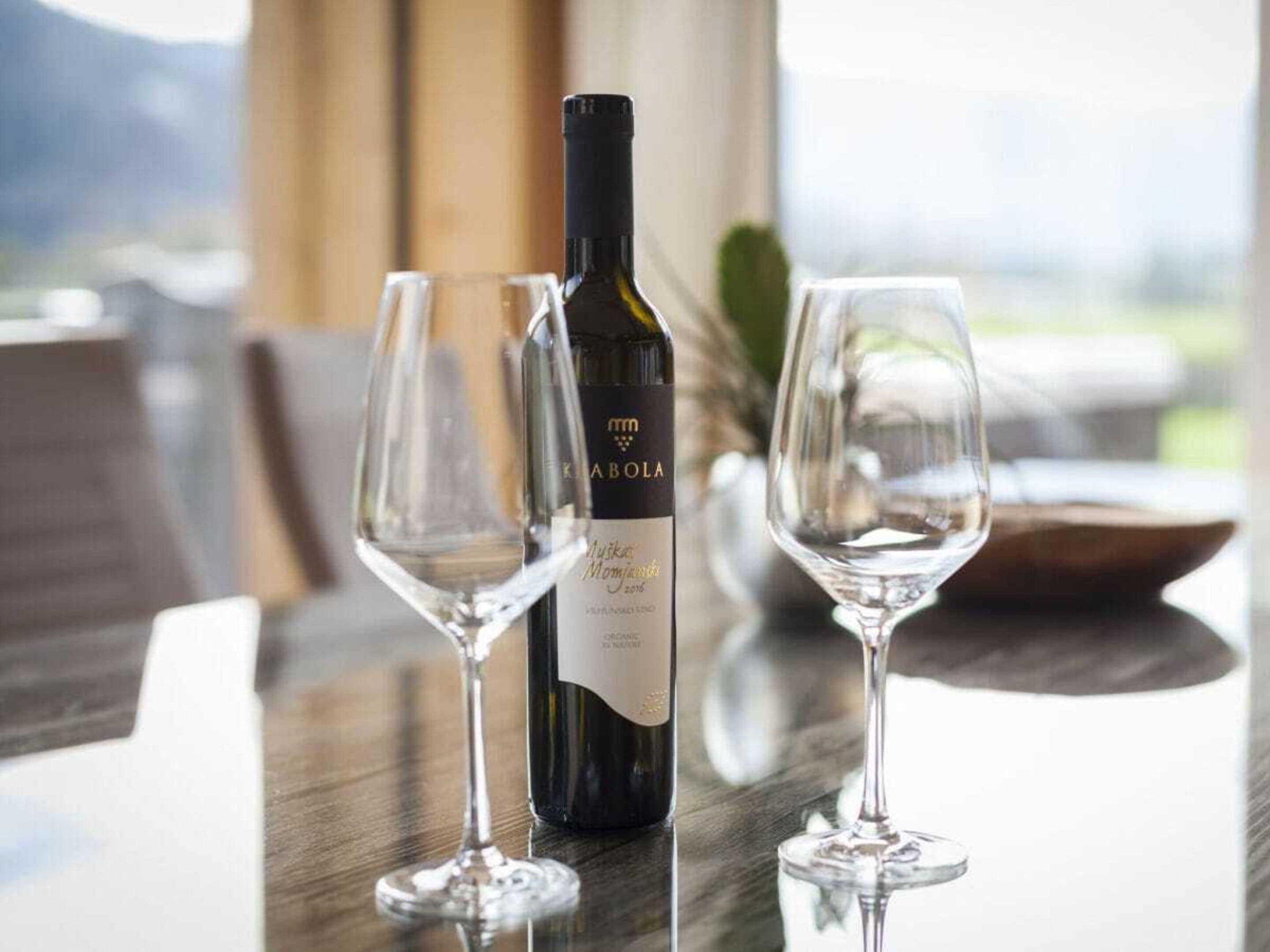 A bottle of wine and two glasses on a table with a view of the outdoors.