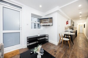 A modern living room with a black TV on the wall, a dining area with a small table and chairs, and a kitchen in the background. - Earl`s Court Residence | Elegant 1BR | London (London)