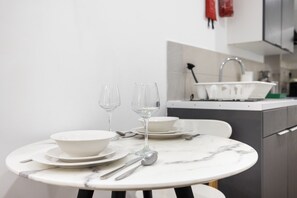 A dining table with white plates, two wine glasses, and cutlery, set against a white wall with a kitchen in the background. - Earl`s Court Residence | Elegant 1BR | London (London)