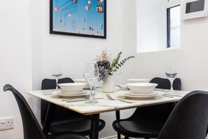 Ein Esstisch ist mit weißen Tellern, Schüsseln und Weingläsern gedeckt, daneben befinden sich schwarze Stühle. Auf der Wand hängt ein Bilderrahmen und ein Blumenstrauß steht in einer Vase.