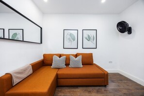 A modern living room with a sectional sofa, framed botanical prints on the wall, and a standing fan. - Earl`s Court Residence | Elegant 1BR | London (London)