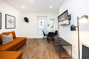 A modern living room with a sofa, a dining table with chairs, a TV mounted on the wall, and a floor lamp. - Earl`s Court Residence | Elegant 1BR | London (London)
